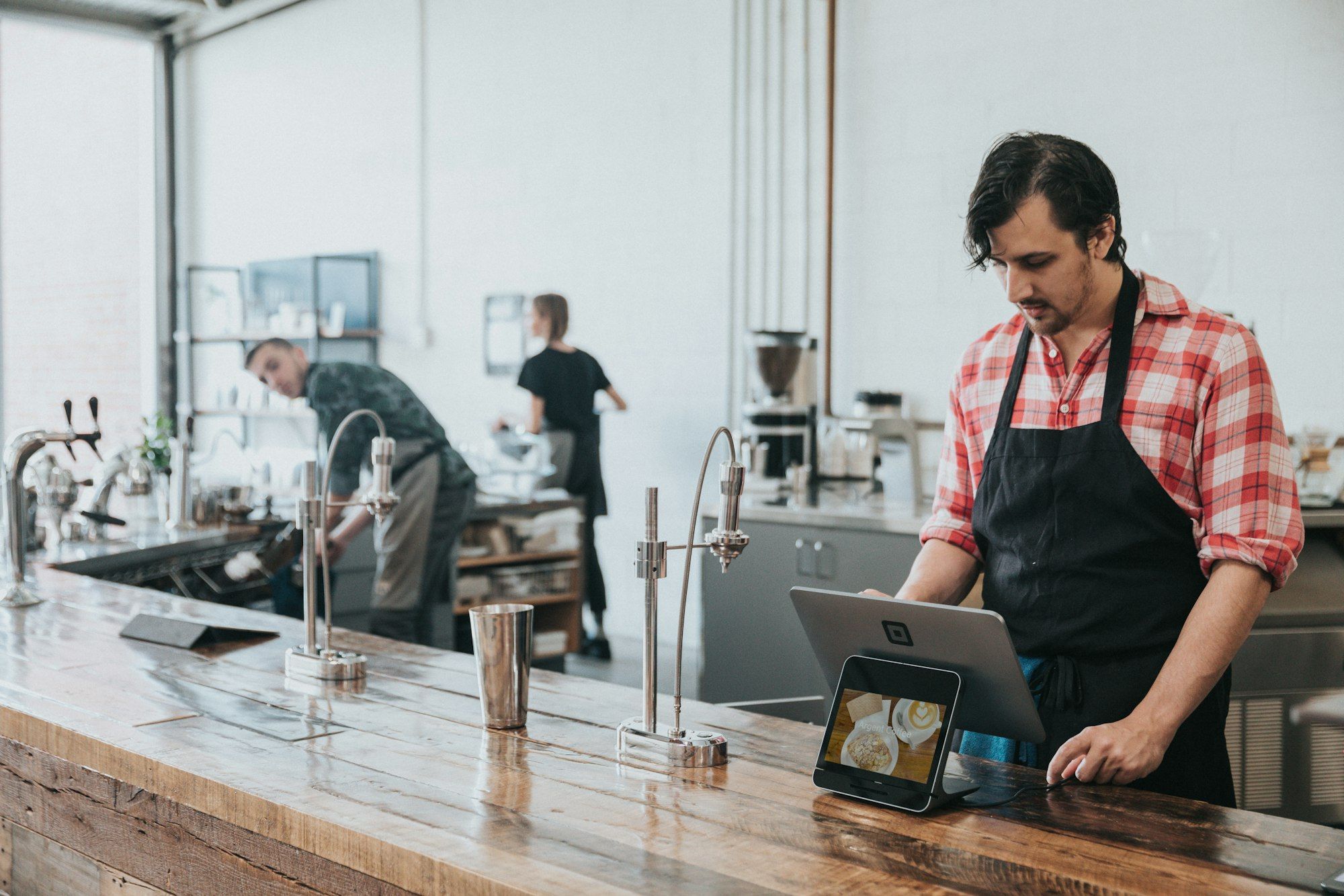 A coffee shop owner in an apron using a tablet at the wooden counter