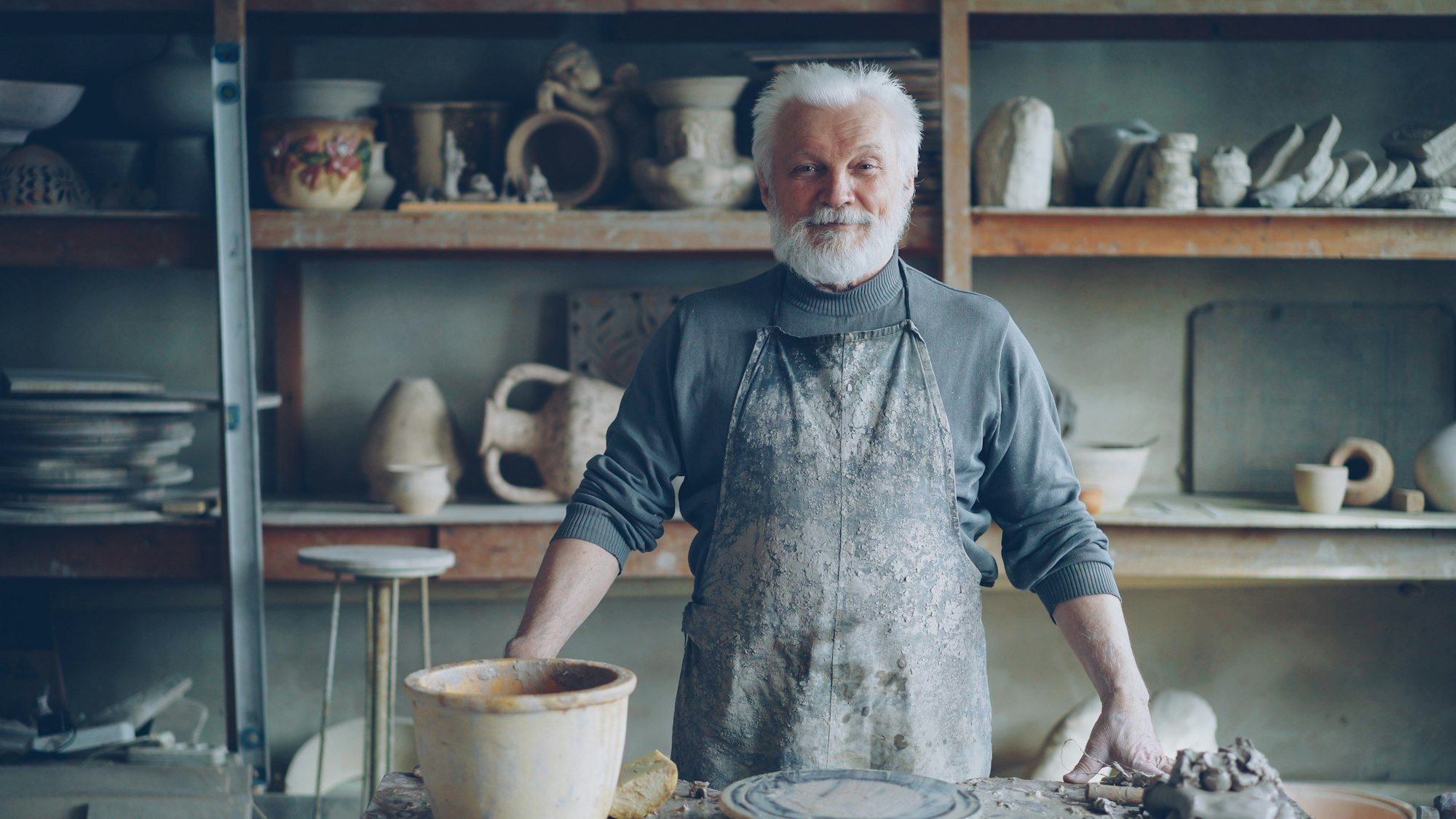 A potter smiling in his workshop surrounded by shelves of finished pieces